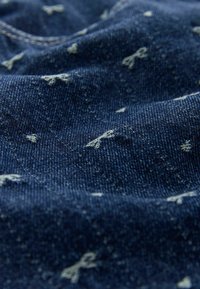 Tamsiai mėlynas džinsinis audinys su šviesiai mėlynomis siuvinėtomis strėlėmis. Jis pasižymi tekstūruotu audimu, minkštu paviršiumi ir subtiliu išblukimu kai kuriose vietose.