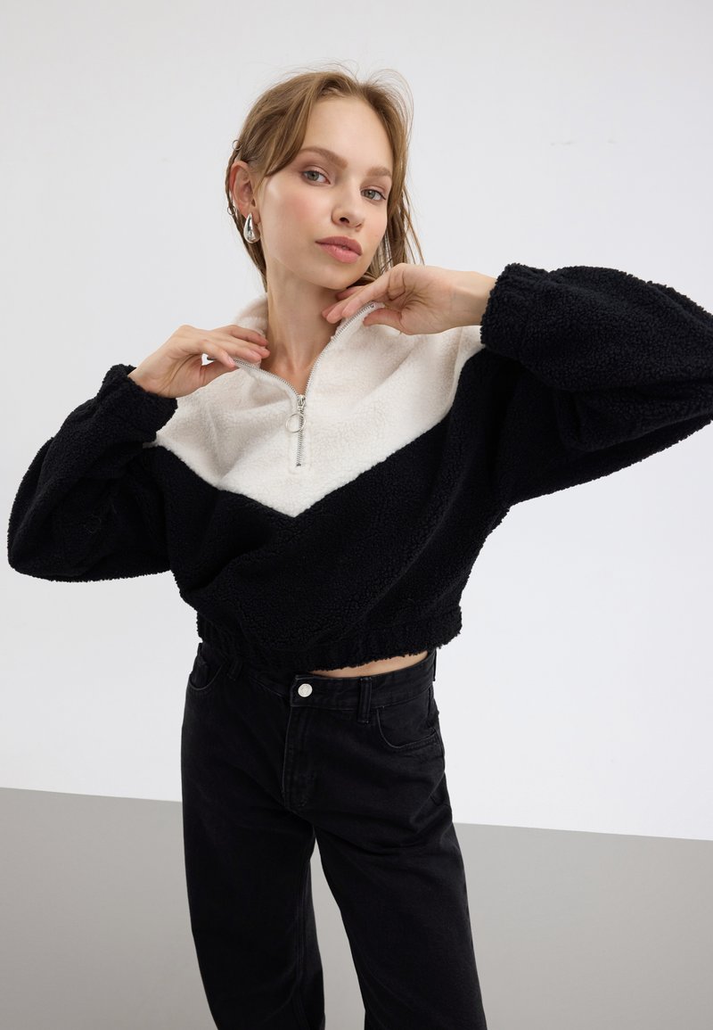Pullover in pile bianco e nero con mezza cerniera, caratterizzato da un design a zigzag e un orlo elastico. Il modello indossa jeans neri e orecchini d'argento.