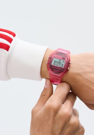 Digitalna ura s prozornim roza rezinastim ohišjem in trakcem. Ima digitalni prikaz, črn številčnik in srebrne poudarke. Vodoodporna.