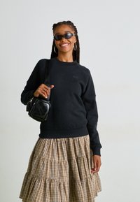 Felpa nera con polsini a costine, abbinata a una gonna a balze a scacchi beige. La modella tiene in mano una piccola borsa a tracolla nera ed indossa occhiali da sole scuri.