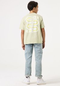 Camiseta verde claro con un diseño gráfico en blanco y amarillo que dice "NUEVA PERSPECTIVA" en la parte de atrás, combinada con jeans azul claro y zapatillas blancas.