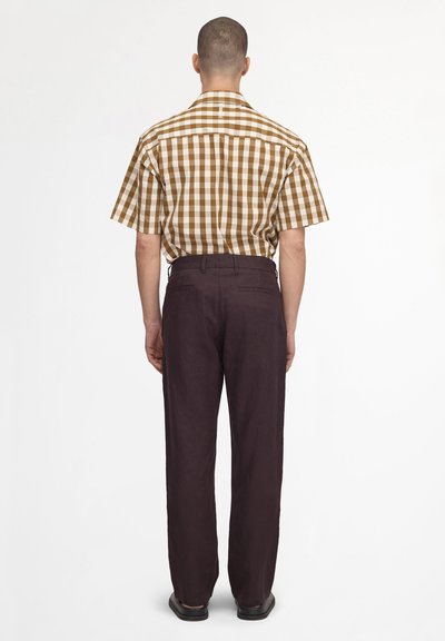 Man standing facing away wearing a brown and white checkered short-sleeve shirt, dark brown pants, and black shoes on a white background.