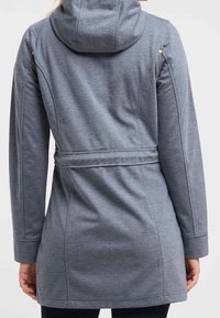 Femme portant un manteau à capuche gris mi-long avec une ceinture, vue de dos, se tenant contre un fond clair.