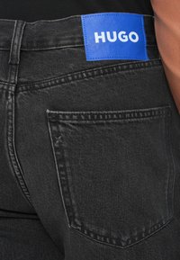 Vaqueros de mezclilla negra con una etiqueta de cuero azul que lleva el texto "HUGO". El diseño incluye un bolsillo trasero y detalles de costura sutiles.