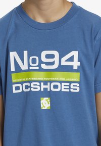 Modrá bavlněná tričko s velkým bílým textem "No 94" a "DC SHOES", se zeleným akcentovým pruhem a logem. Kulatý výstřih, volný střih.