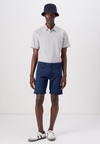 Chemise à manches courtes rayée, short bleu marine, baskets gris et blanc, et un chapeau de seau noir. Tissu en coton, coupe décontractée, ourlet roulé sur le short.