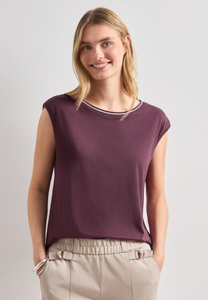 Femme souriante aux cheveux blonds mi-longs, portant un haut sans manches violet foncé et un pantalon beige clair, les mains dans les poches.