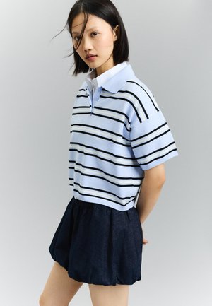 Kvinde med glat sort hår iført en lyseblå, kortærmet poloshirt med striber og en mørkeblå ballonnederdel, stående foran en grå baggrund.