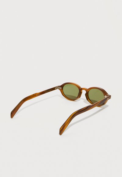 Lunettes de soleil rondes avec des verres verts, monture écaille de tortue marron et branches courbées reposant sur une surface blanche.