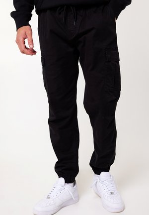 Black cargo pants with elastic cuffs and drawstring waistband, featuring side pockets and a relaxed fit, paired with white sneakers.