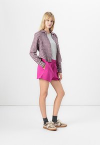 Ung kvinna står med händerna i fickorna, iklädd en rutiga skjorta, grå t-shirt, ljusrosa shorts, mörka strumpor och sneakers.