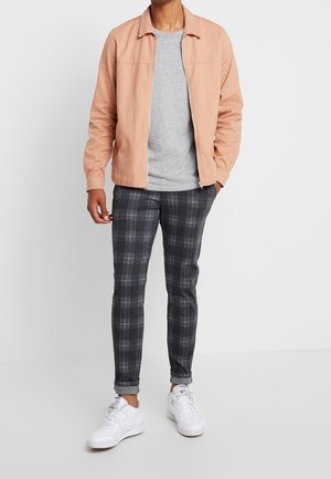 Licht perzikkleurige zip-up jas, grijze t-shirt, zwart geruite strakzittende broek en witte sneakers. Focus op casual stijl met contrasterende kleuren en patronen.