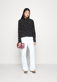 Blusa preta de manga longa com padrão de folhas multicoloridas, combinada com calças largas brancas. O modelo segura uma carteira de mão rosa brilhante.