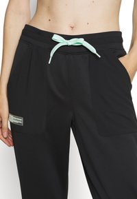 Pantalones deportivos negros con una textura suave, que cuentan con un cordón de color verde menta y bolsillos laterales. Incluye una etiqueta en el muslo izquierdo.
