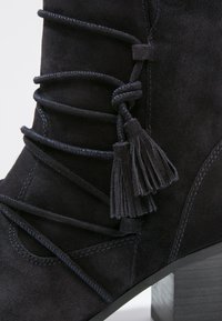 Bottines en suède noir avec des lacets décoratifs et un accent de pompon, dotées d'un talon empilé et de détails de couture contrastants.