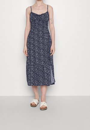 Abito midi floreale blu navy con spalline sottili, corpetto aderente e gonna svasata. Presenta una leggera apertura e tessuto testurizzato. Abbinato a sandali bianchi.
