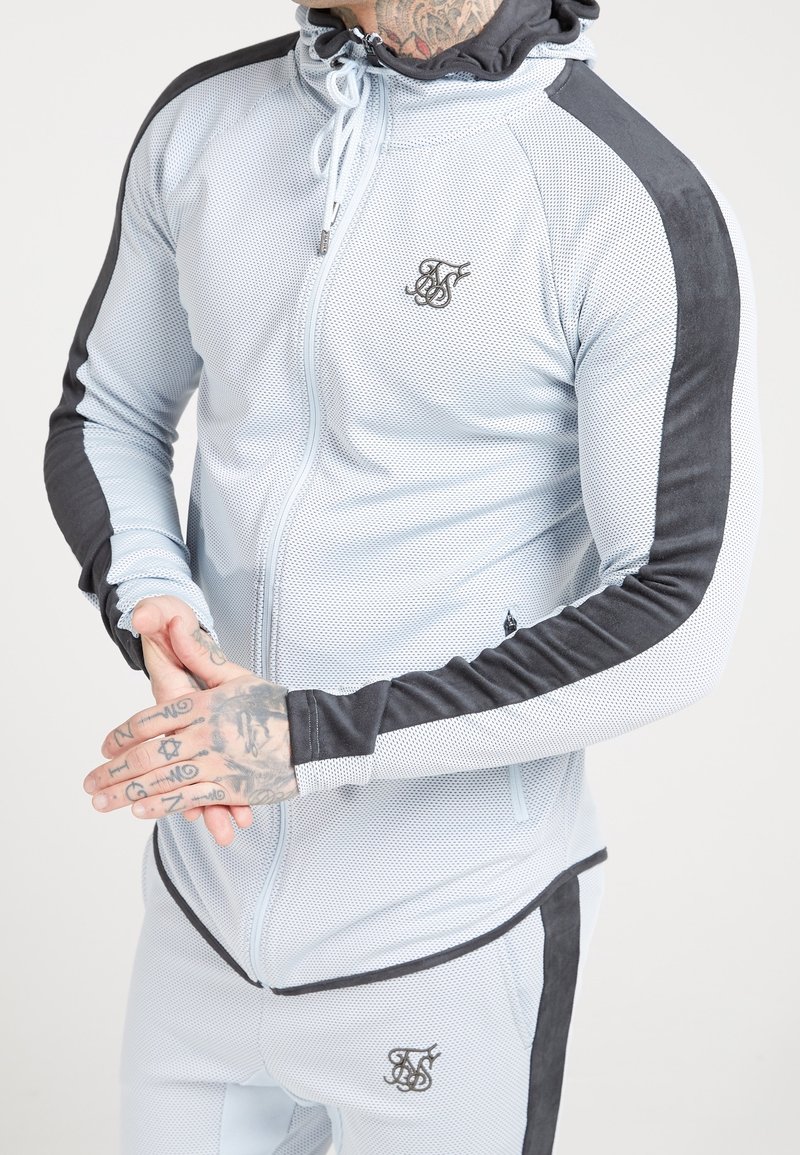 Lyseblå mesh zip-up jakke med sorte ruskindsdetaljer; har hætte, snor og logo på brystet; tætsiddende design og struktureret stof.