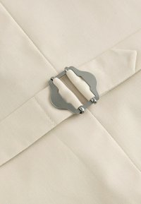 Beige stof met een gladde textuur, met een zilveren metalen gesp en twee stoffen banden, aangebracht op een gevouwen gedeelte.