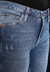 Jeans de mezclilla color azul claro con una textura desgastada, costuras detalladas, cierre con botón y un diseño tradicional de cinco bolsillos.