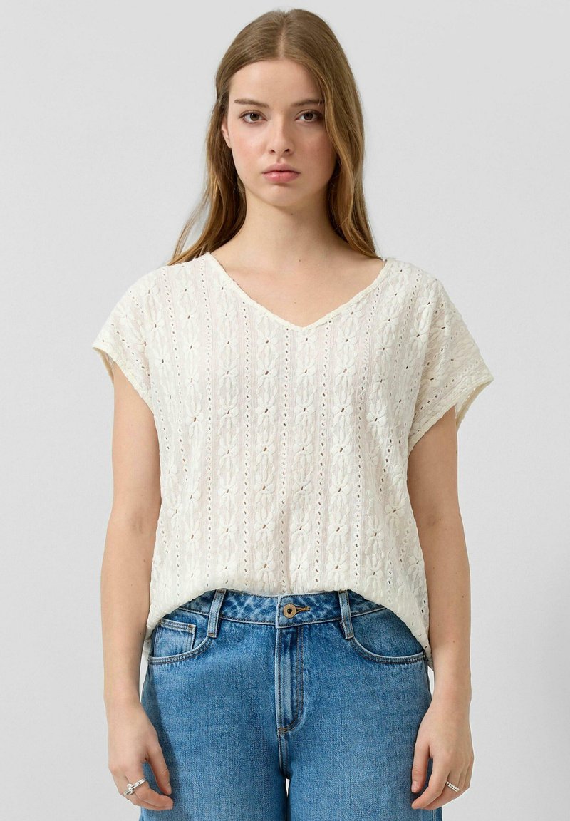 Joven mujer vestida con una blusa blanca de encaje de manga corta y jeans azules de cintura alta, posando frente a un fondo liso.