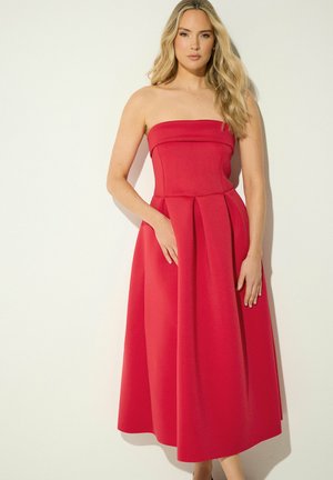 Frau mit langen blonden Haaren, die ein trägerloses, plissiertes rotes Midikleid trägt, steht vor einem schlichten hellen Hintergrund.