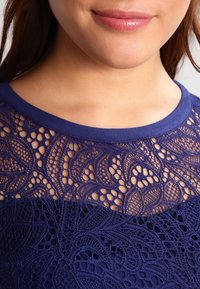 Top blu navy con dettagli in pizzo intricati sulla parte superiore, con un tessuto solido e morbido sulla parte inferiore. Collo rotondo.