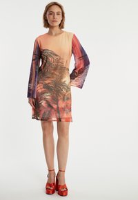 Robe longue en voile à manches longues avec un dégradé orange-violet, présentant un motif de palmiers tropicaux et une coupe ample. Une paire de sandales à plateforme rouges est visible.