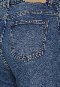 Jeans de denim com um desbotado azul, apresentando um pequeno bolso traseiro em forma de diamante e costura visível. Uma etiqueta de couro bege está presa na cintura.