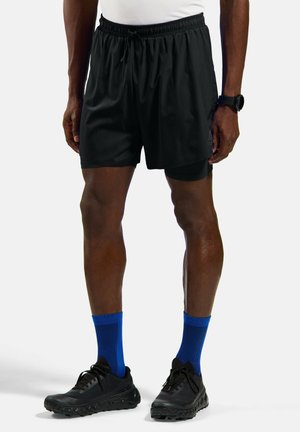 Man draagt zwarte sportshorts met ingebouwde compressielaag, blauwe crew-sokken, zwarte hardloopschoenen, wit shirt en zwarte polshorloge.