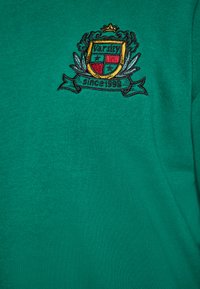 Groene katoenen sweatshirt met een geborduurde embleem en de tekst "Varsity" en "sinds 1998", afgewerkt met gouden en zwarte accenten.