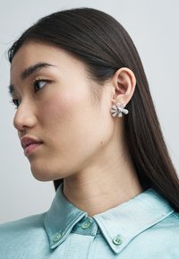 Sidabrinės gėlės formos auskaras su tekstūruotais žiedlapiais ir centrine puošmena, dėvimas žmogaus, kuris vilki šviesiai mėlyną satino marškinius su sagomis.