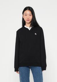 Sort sweatshirt med V-hals, lange ærmer og en hvid indvendig krave. Har et lille logo-detail på brystet. Bæres med blå jeans.