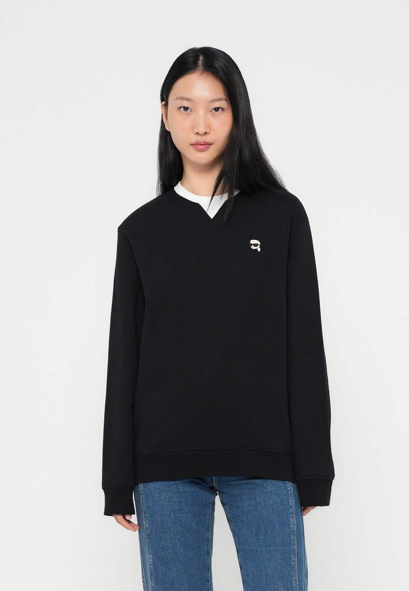 Sweatshirt noir avec un col en V, des manches longues et un col intérieur blanc. Présente un petit détail de logo sur la poitrine. Porté avec un jean bleu.