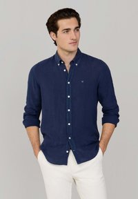 Jonge man draagt een marineblauw overhemd met opgerolde mouwen en witte broek, staat met handen in de zakken en kijkt opzij.