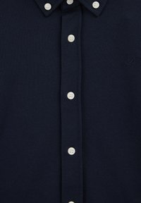 Tumši zilas pogas krekls ar baltām pogām un smalki izšūtu putna logo uz kreisā krūšu. 