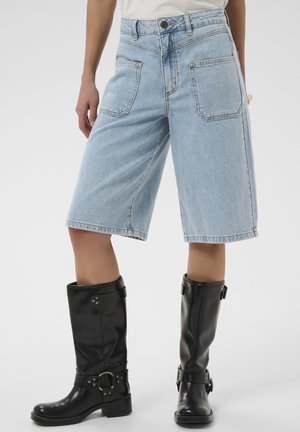 Personne portant un short en jean bleu clair jusqu'aux genoux et des bottes en cuir noires mi-mollet avec des détails de boucle, debout sur un fond uni.