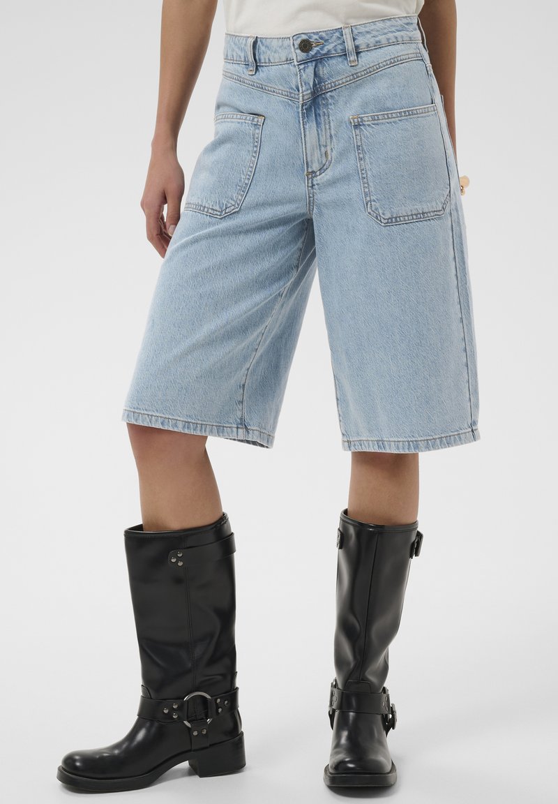 Personne portant un short en jean bleu clair jusqu'aux genoux et des bottes en cuir noires mi-mollet avec des détails de boucle, debout sur un fond uni.