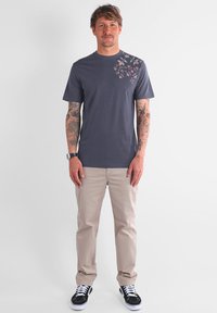 T-shirt en coton gris avec un imprimé floral sur l'épaule gauche, associé à un pantalon beige et des baskets noires. Homme debout, les bras détendus.
