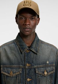 Jeansjacka med en urblekt blå finish, med två bröstfickor och kopparfärgad hårdvara. Modellen bär en khaki-keps med "HUGO"-logotyp.
