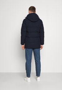 Hombre de pie de espaldas, vistiendo una chaqueta de plumas con capucha negra, jeans azules y zapatillas blancas, frente a un fondo liso.