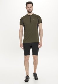 Olivgrünes kurzärmeliges Fahrradtrikot mit einem halben Reißverschluss, schwarzen Akzenten, kombiniert mit schwarzen eng anliegenden Shorts und schwarzen Fahrradschuhen.