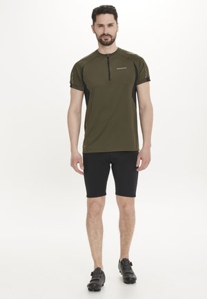 Olivgrünes kurzärmeliges Fahrradtrikot mit einem halben Reißverschluss, schwarzen Akzenten, kombiniert mit schwarzen eng anliegenden Shorts und schwarzen Fahrradschuhen.