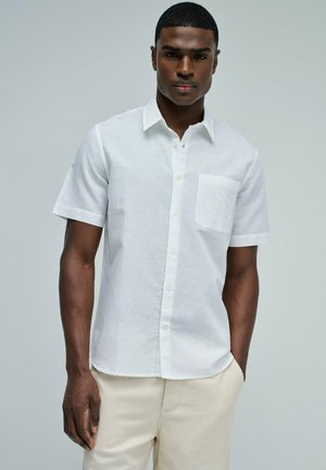 Hombre con camisa blanca de manga corta con bolsillo en el pecho y pantalones beiges claros, de pie frente a un fondo gris liso.