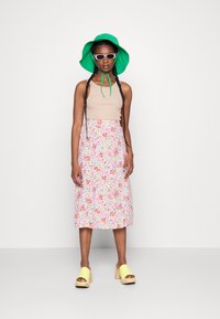 Camisola bege de alças, saia midi floral com padrões em rosa, vermelho e branco, chapéu de balde verde, óculos de sol brancos e tamancos amarelos com solas de madeira.