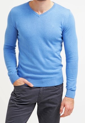 Homme portant un pull à col en V bleu clair et un pantalon gris foncé, une main dans la poche et l'autre reposant à côté.
