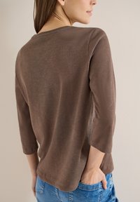 Femme portant une chemise marron à manches longues et un jean bleu, debout avec une main dans sa poche, tournée dos à la caméra.