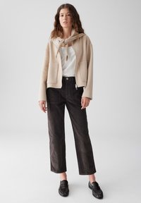 Donna che indossa una giacca beige con zip, un maglione annodato attorno al collo, una maglietta bianca, pantaloni neri e mocassini neri, in piedi su uno sfondo chiaro uniforme.