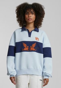 Lys blå oversized sweatshirt med mørkeblå horisontale striper, har broderte detaljer med oransje aksenter. Knappekrage.