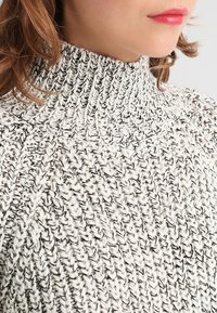 Stickad tröja i svart och vitt garn med hög krage, texturerat mönster och synliga sömnadsdetaljer. Inga ytterligare dekorationer.