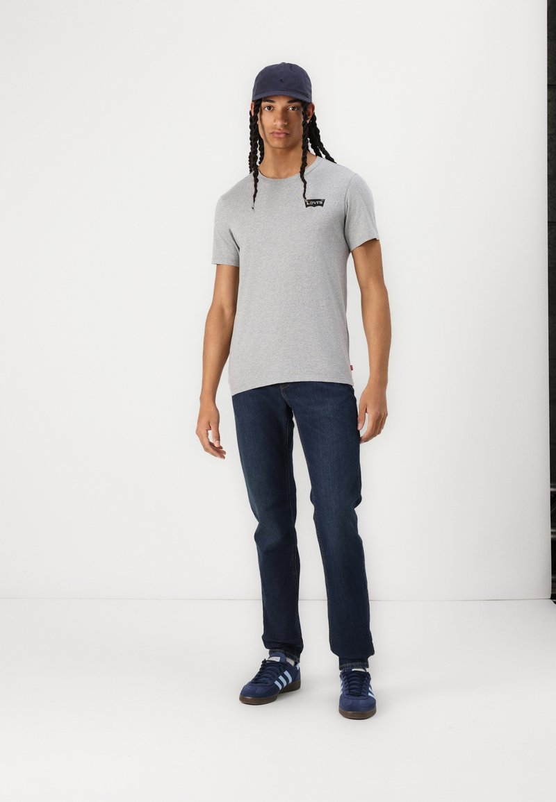 Camiseta de algodón gris con un pequeño logo negro, jeans azul oscuro y zapatillas azules con rayas azul claro, combinada con una gorra marina.